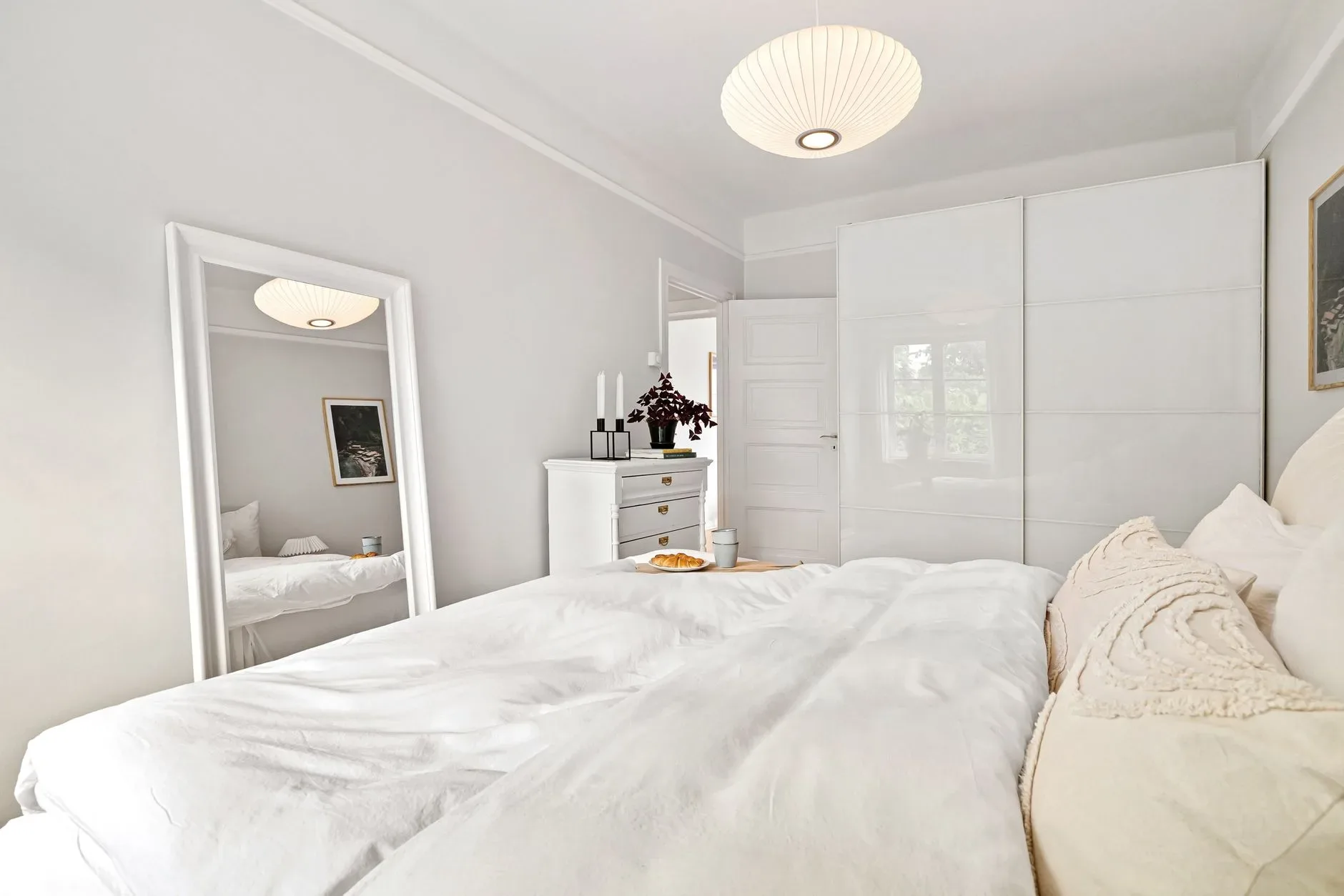 A minimalist bedroom with white decor, featuring a bed, dresser, and mirror under soft lighting.