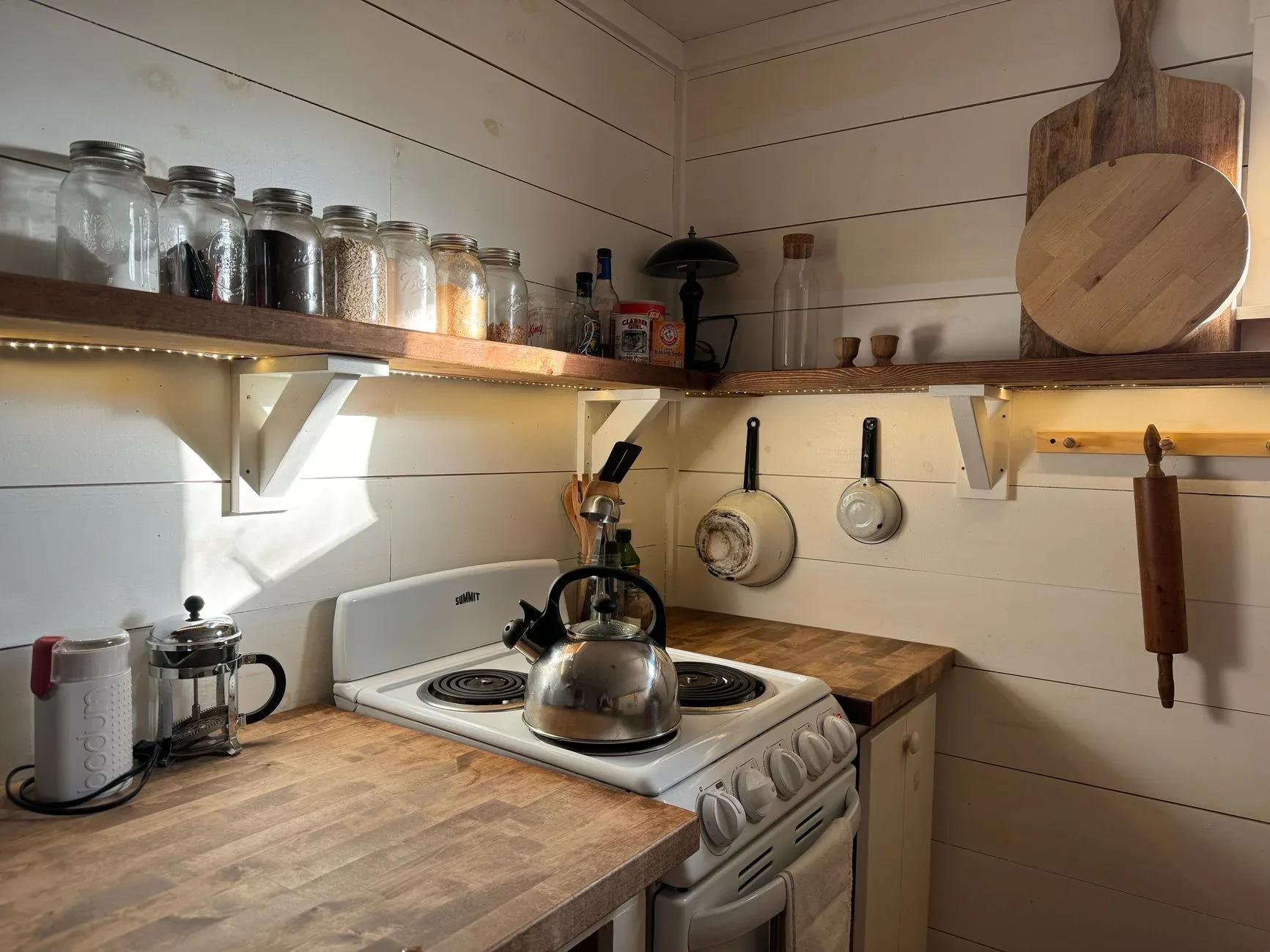 A small rustic kitchen with a wooden shelf, stove, and cooking utensils, offering a warm and cozy atmosphere.