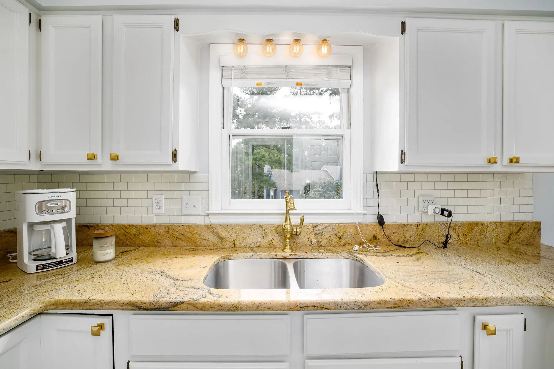 Bright kitchen with a marble countertop, golden faucet, and window view. Features a coffee maker and subway tile backsplash.