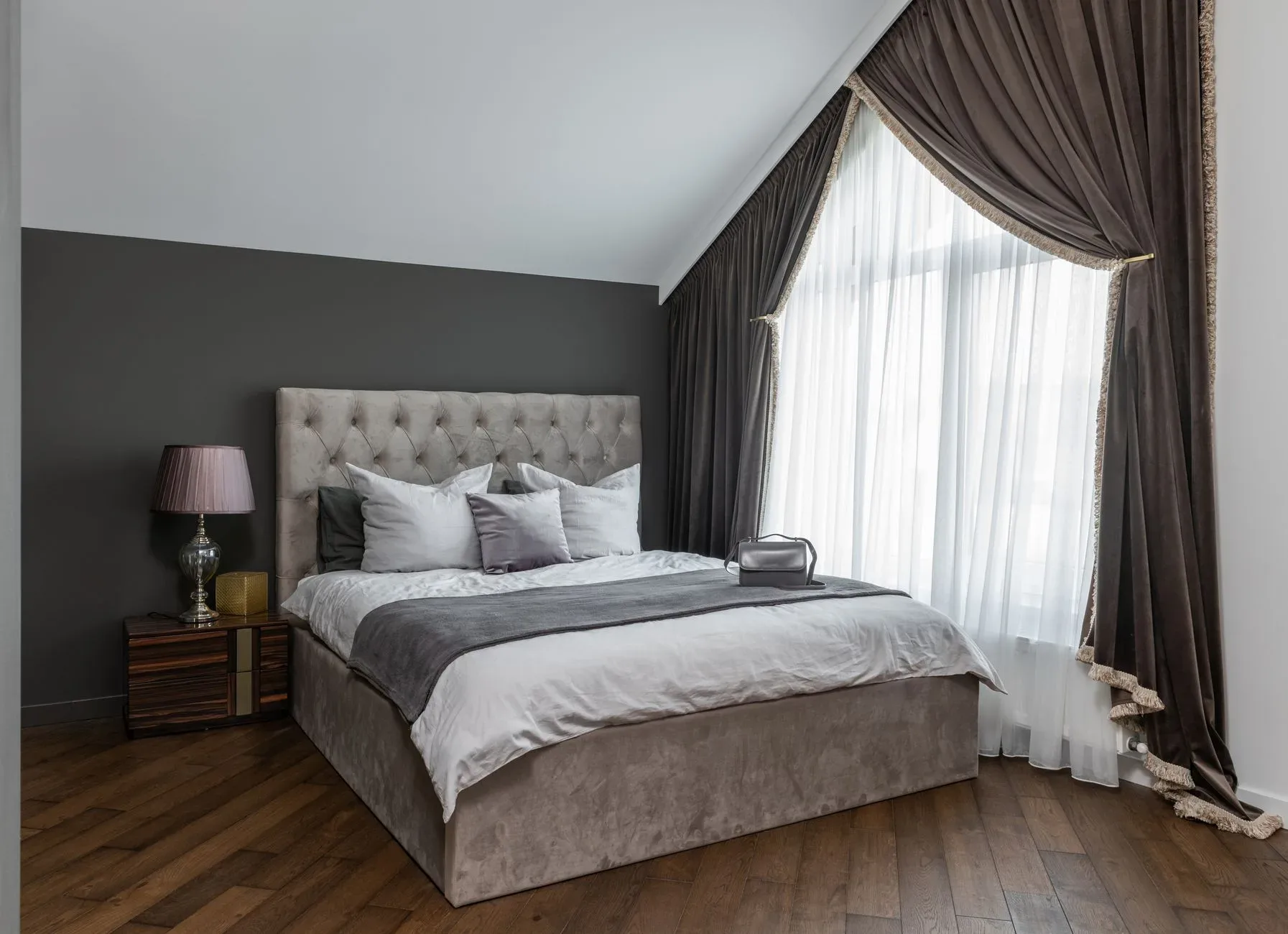 Comfortable bed with blanket and cushions placed on parquet near window with curtains and lamp on bedside table in modern bedroom