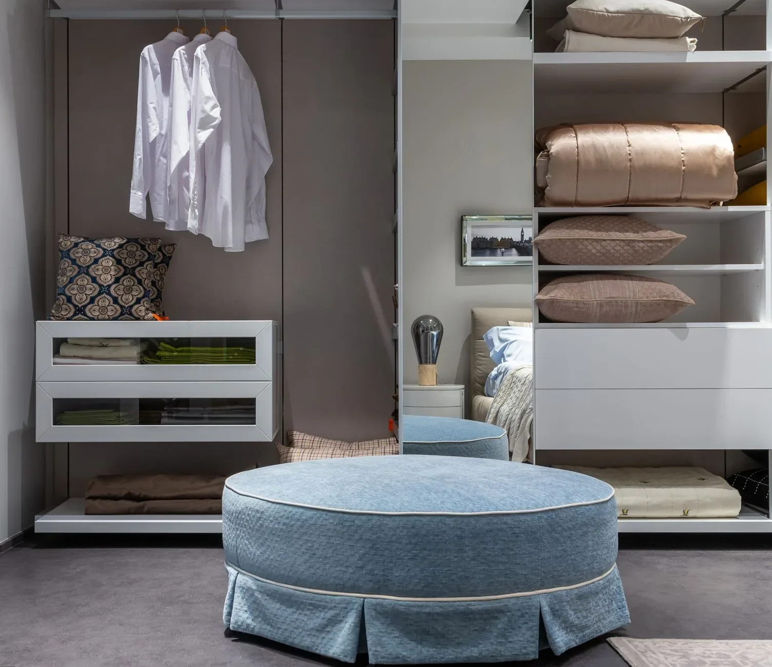 Comfortable round pouf near opened wardrobe with hanging clothes and bedding on shelves placed against mirror with reflection if bed in bedroom