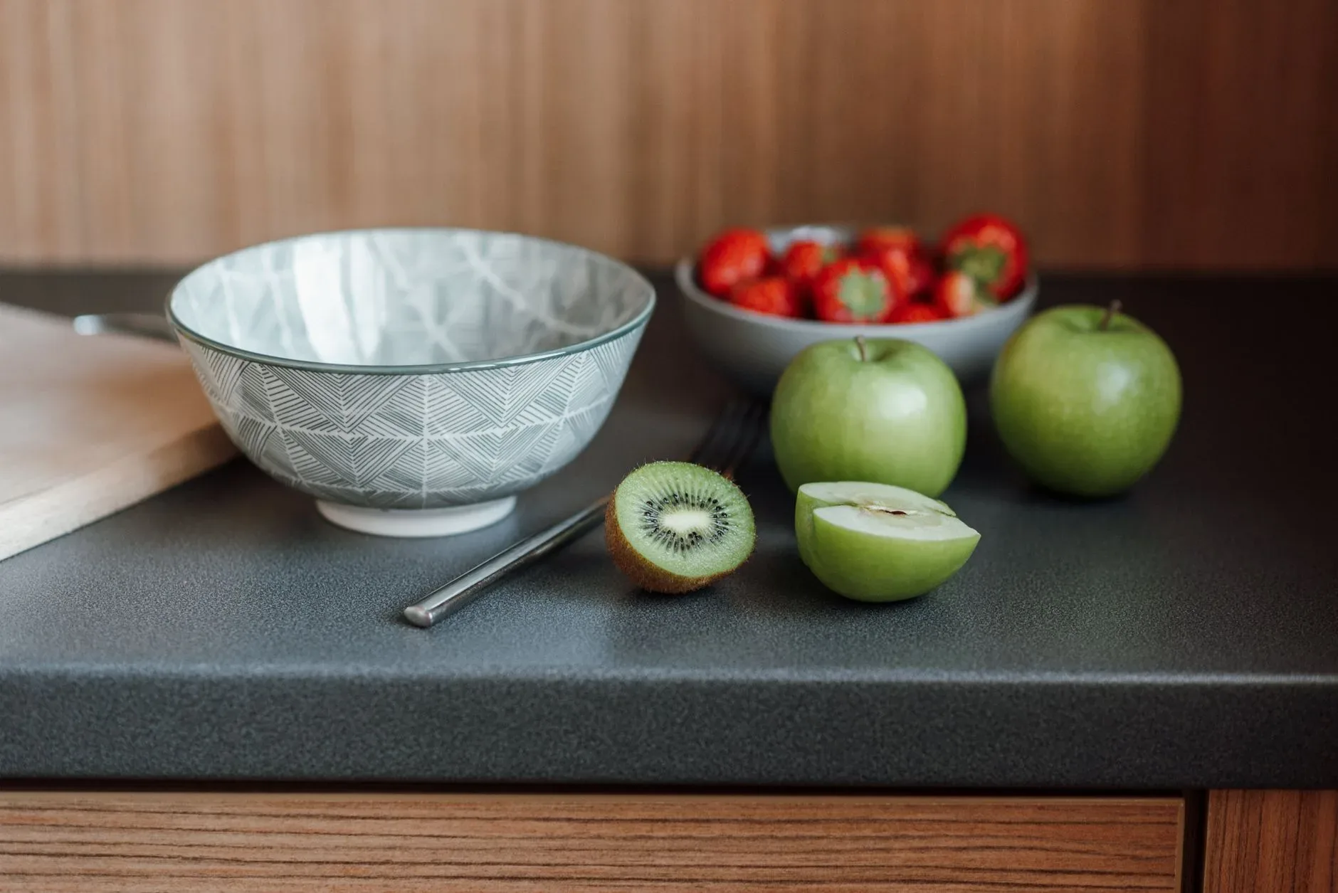 Table with bowl near fork and green apples with kiwi and cutting board near strawberries in dishware in light kitchen