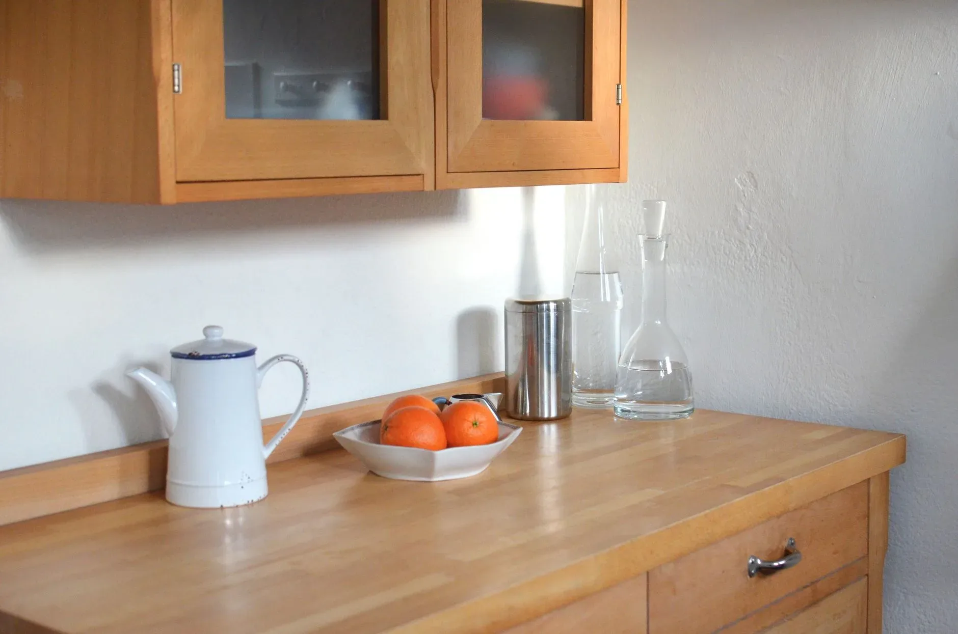 Wooden cabinet for dishes placed above wooden table with teapot glassware and bowl of oranges in modern light kitchen